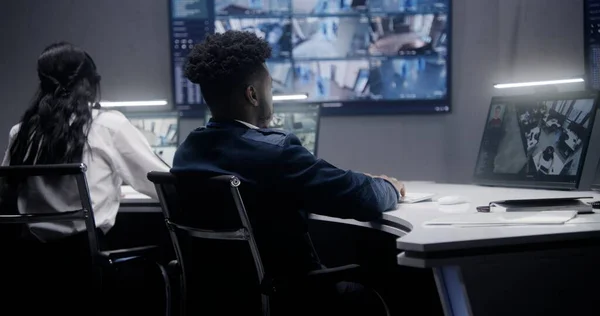 非裔美国人安全操作员用人工智能人脸识别监控闭路电视摄像机 墙上的多个大屏幕显示了摄像头的视图 同事在后台工作 社会安全概念 — 图库照片