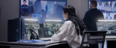 Bayan güvenlik görevlisi polis izleme merkezinde bilgisayar üzerinde çalışıyor. Çoklu etnik işçiler güvenlik kameralarını kontrol ediyor. Yapay zeka tablet ve büyük dijital ekranda tanıma sistemiyle karşı karşıya..