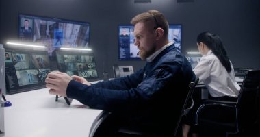 Erkek güvenlik görevlisi tablete dokunuyor ve yapay zeka yüz taramasıyla mobese kameralarını kontrol ediyor. Bilgisayar monitörleri ve güvenlik kamerası görüntülerini gösteren büyük dijital ekranlar. Bayan meslektaşım arka planda çalışıyor..
