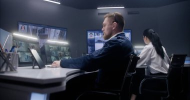 Erkek güvenlik görevlisi tablete dokunuyor ve yapay zeka yüz taramasıyla mobese kameralarını kontrol ediyor. Bilgisayar monitörleri ve güvenlik kamerası görüntülerini gösteren büyük dijital ekranlar. Bayan meslektaşım arka planda çalışıyor..