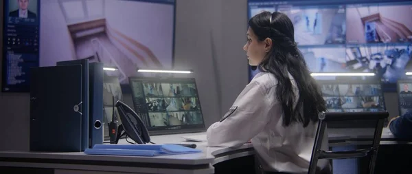 Female security officer works on computer in police monitoring center. Multi ethnic workers control surveillance CCTV cameras with tracking AI face recognition system on tablet and big digital screen.