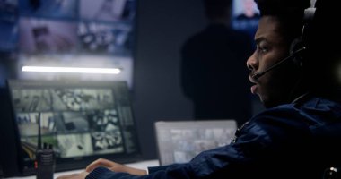Kulaklıklı Afro-Amerikan polis memuru, güvenlik kameralarını kontrol ediyor. Yapay zeka, polis izleme odasında tarama yapıyor. Arka planda CCTV görüntüsü olan bir sürü büyük ekran. Sosyal güvenlik kavramı. Kapat..