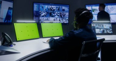 Karışık yarış memurları, operatörler güvenlik kameralarını kontrol ediyor. Çalışanlar izleme merkezinde çalışıyor. Güvenlik kameralarının görüntülerini gösteren büyük dijital ekranlar. Yeşil ekranlı bilgisayar monitörleri.