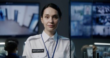Polis gözetleme merkezindeki kadın güvenlik görevlisi kameraya bakıyor. Erkek çalışanlar arka planda çalışır. CCTV kameraları büyük dijital ekranda gösteriliyor. İzleme ve izleme sistemi. Portre.