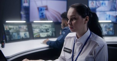 Afrikalı Amerikalı erkek güvenlik operatörü polis gözlem merkezinde meslektaşıyla telefonla konuşuyor. Kadın kamera görüntüsü olan monitörlere bakıyor. Duvardaki büyük ekranlar kamera görüntülerini gösteriyor.