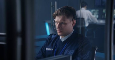 Kulaklıktaki güvenlik görevlisi güvenlik kameralarını kontrol ediyor. Güvenlik izleme merkezinin erkek çalışanı. Meslektaşlar ve arka planda güvenlik kameraları olan büyük dijital ekranlar..