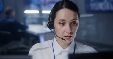 Kulaklıktaki kadın güvenlik operatörü, bilgisayardaki CCTV kameralarını kontrol ediyor. Güvenlik Gözlem Merkezi çalışanı. Meslektaşlar ve arka planda güvenlik kameraları olan büyük dijital ekranlar..