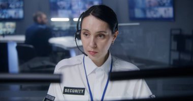 Kulaklıktaki kadın güvenlik operatörü, bilgisayardaki CCTV kameralarını kontrol ediyor. Güvenlik Gözlem Merkezi çalışanı. Meslektaşlar ve arka planda güvenlik kameraları olan büyük dijital ekranlar..