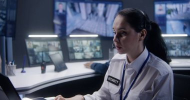 Afrikalı Amerikalı erkek güvenlik operatörü polis gözlem merkezinde meslektaşıyla telefonla konuşuyor. Kadın kamera görüntüsü olan monitörlere bakıyor. Duvardaki büyük ekranlar kamera görüntülerini gösteriyor.