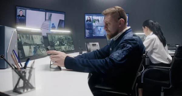在警察监控中心 耳机中的男性安全操作员用人工智能面部识别来控制闭路电视摄像机 平板电脑 电脑显示器和显示安全摄像头镜头的大型数字屏幕 — 图库照片