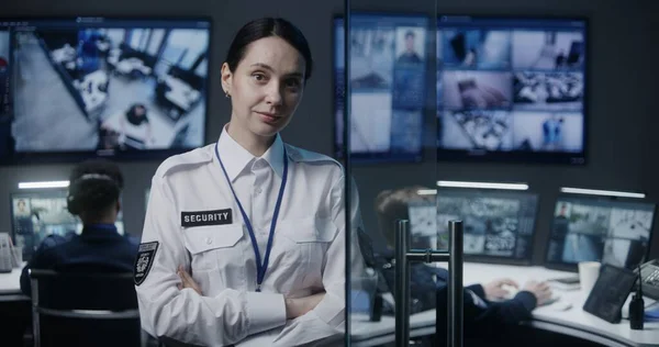 Female security worker in police surveillance center looks at camera. Male employees work on background. CCTV cameras footage displayed on big digital screen. Tracking and monitoring system. Portrait.