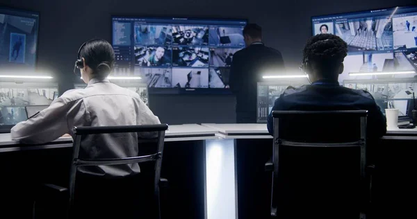 Multi ethnic security employees monitor CCTV cameras in police surveillance center. Multiple big screens on the wall and PC monitors showing security cameras footage. Tracking system. Social safety.