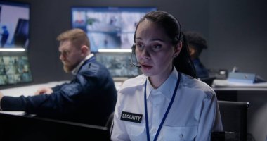 Kulaklıklı kadın güvenlik operatörünün polis gözetleme merkezindeki kamera görüntülerine yakından bakın. Arka planda çok ırklı meslektaşlar çalışıyor. Sosyal güvenlik kavramı.