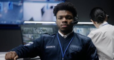 Polis izleme merkezindeki Afro-Amerikan güvenlik görevlisi gülümseyip kameraya bakıyor. Kadın operatör bilgisayardaki CCTV kameralarını kontrol ediyor. Kameralı büyük dijital ekran..