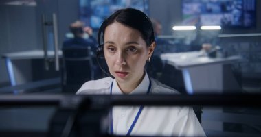 Kulaklıktaki kadın güvenlik operatörü, bilgisayardaki CCTV kameralarını kontrol ediyor. Güvenlik Gözlem Merkezi çalışanı. Meslektaşlar ve arka planda güvenlik kameraları olan büyük dijital ekranlar..