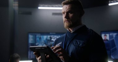 Polis gözetleme merkezindeki güvenlik kameralarını kontrol eden erkek güvenlik görevlisi tablet kullanıyor. Duvardaki büyük ekranlar güvenlik kamerası görüntülerini gösteriyor. İzliyorum. Sosyal güvenlik kavramı. Portre.