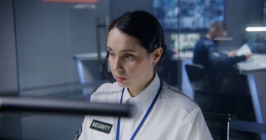 Kulaklıktaki kadın güvenlik operatörü, bilgisayardaki CCTV kameralarını kontrol ediyor. Güvenlik Gözlem Merkezi çalışanı. Meslektaşlar ve arka planda güvenlik kameraları olan büyük dijital ekranlar..