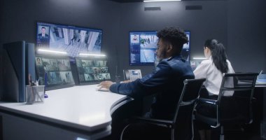 Çoklu etnik güvenlik operatörleri, polis gözetleme merkezindeki yapay zeka yüz taramasıyla CCTV kameralarını kontrol ediyor. Afrikalı Amerikalı subay telefonla konuşuyor. Duvardaki ekranlar güvenlik kamerası görüntülerini gösteriyor..