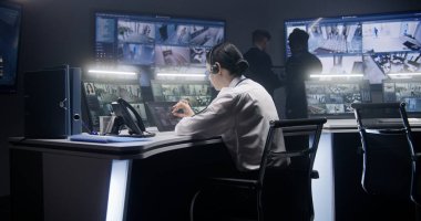Kulaklık takan kadın güvenlik görevlisi tablet kullanarak kameraları kontrol ediyor. Meslektaşlar arka planda çalışır. Duvarda CCTV görüntülerini gösteren büyük ekranlar var. İzliyorum. Sosyal güvenlik kavramı