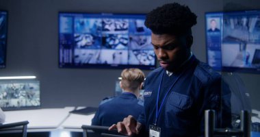 Afro-Amerikan erkek subay tablet kullanıyor. Güvenlik kontrol merkezinde çok ırklı çalışanlar. Büyük dijital ekranlar ve güvenlik kameralı bilgisayar monitörleri..