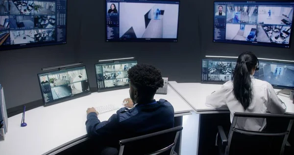 Multi ethnic security officers control CCTV cameras with AI facial scanning in police monitoring center. African American operator talks by phone. Screens on the wall showing security cameras footage.