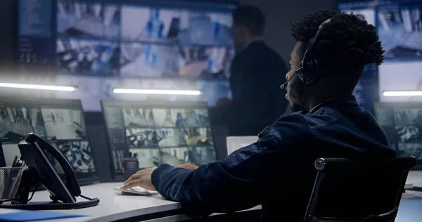 戴耳机的非裔美国男性操作员在警察监视室内用人工智能面部扫描来监控摄像头 多个大屏幕 背景上有闭路电视录像 社会安全的概念 — 图库照片