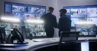 Çok ırklı çalışanlar konuşuyor ve CCTV kameralarını kontrol ediyor. Yapay zeka da izleme merkezinde birlikte tarama yapıyor. Duvarda güvenlik kamerası görüntülerini gösteren büyük ekranlar var. Sosyal güvenlik kavramı.