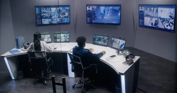 Multi ethnic security officers monitor CCTV cameras with AI face scanning. Multiple big screens with displayed security cameras. Teamwork in surveillance room. Monitoring, tracking and social safety.