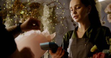 Kadın çiçekçi müşteriye ödeme için terminal veriyor. Çiçekçi dükkanındaki akıllı saatleri kullanarak bir buket çiçeğin parasını temassız ödemeyle ödüyor. Çiçekçilik, küçük işletme konsepti.