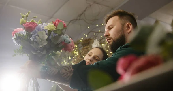 Two entrepreneurs florists together create beautiful bouquet in flowers shop. Man adds fresh flowers to bunch. Woman looks on flower arrangement. Retail floral business and entrepreneurship concept.