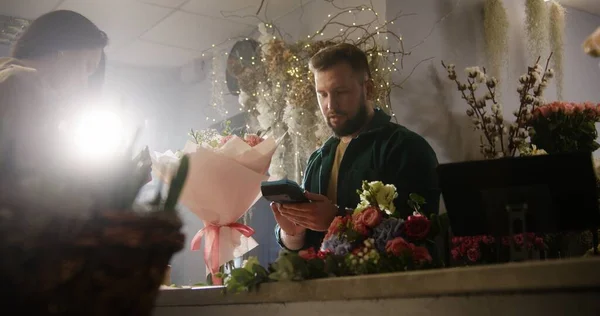 Woman buys bouquet in flower shop and pays for purchase with contactless payment using smartphone. Male florist sells fresh flowers to female customer. Floristry, retail floral small business concept.