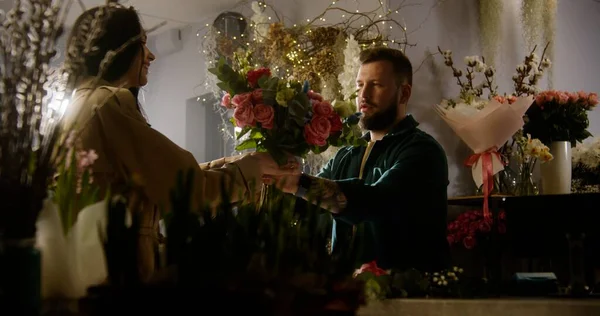 Woman buys bouquet in flower shop and pays for purchase with contactless payment using phone. Male florist sells fresh flowers to female customer. Concept of floristry, retail floral small business.