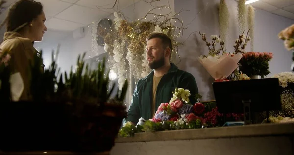 Male florist, seller gives fresh flowers to the female buyer and talks with her. Woman buys spring bouquet in flower shop. Concept of floristry, retail floral small business and entrepreneurship.
