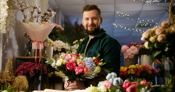Professional florist collects and keeps beautiful bouquet in hands, smiles and looks at camera. Vases with fresh flowers stand behind man. Retail floral business and entrepreneurship concept. Portrait