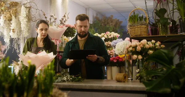 Florist counts fresh flowers in vase and takes customer order online using digital tablet computer. Female colleague wraps bouquet with wrapping paper. Concept of floral business and entrepreneurship.