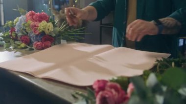 Profesyonel bir çiçekçi masa örtüsü kağıdı koyar ve makasla keser. Çiçekçide çalışma süreci. Yakınlarda güzel bir buket var. Perakende satış işi ve girişimcilik kavramı.