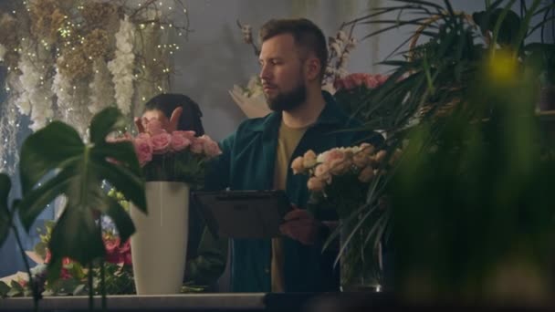 男性花屋は花瓶に花をカウントし デジタルタブレットコンピュータを使用して花店でオンライン注文を取ります 女性同僚が花束を集める 小売業の花小企業と起業家精神 — ストック動画