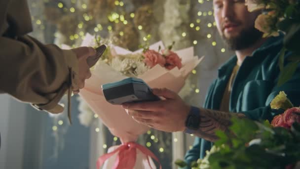男性花屋は女性の顧客への支払いのための端末を提供します 女性は花の店で電話を使用して非接触支払で包装紙に花の花束を支払う フロリストリー 小規模ビジネスコンセプト — ストック動画