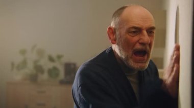 Sinirlenmiş, sinirli yaşlı ve yetişkin bir adam elleriyle duvara vurup ince duvarların ardından birbirine bağırıyor. Komşular kavga eder. Ses yalıtımı olmayan bir daire. Mahalle ve yaşam tarzı kavramı.