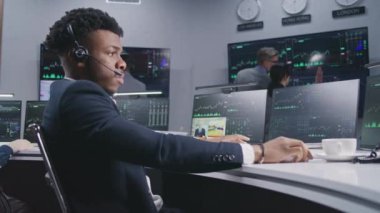 Afro-Amerikalı erkek tüccar bilgisayarda gerçek zamanlı sergilenen hisselerle çalışıyor. Meslektaşlar, arka plandaki birçok büyük ekrandaki döviz piyasası grafiklerini analiz ederler. Kripto para birimi ticareti ve yatırımı.