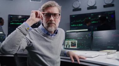 Olgun bir finans analisti gerçek zamanlı hisse senetleri ile PC 'de çalışır, döner ve kameraya bakar. Meslektaşlar, arka plandaki büyük ekranlardaki döviz piyasalarının grafiklerini analiz ederler. Yatırım konsepti. Portre.