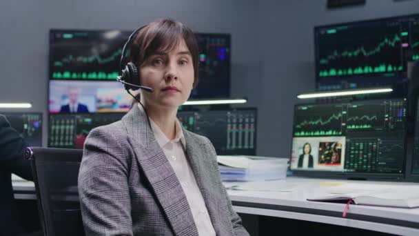 ヘッドセットの女性トレーダーは 表示されたリアルタイム株式とコンピュータに座って カメラを見ています 同僚は バックグラウンドで大きな画面上の取引所市場チャートを分析します 投資と取引 肖像画 — ストック動画