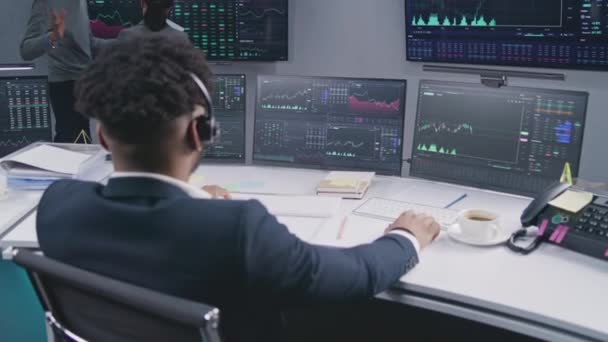 アフリカ系アメリカ人のトレーダーは 表示されたリアルタイム株式を持つコンピュータで動作します 同僚は バックグラウンドで大きな画面上の取引所市場チャートを分析します 取引と投資の概念 バックビュー ドリーショット — ストック動画