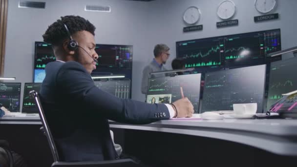 非裔美国商人在电脑上做笔记和工作 并展示实时股票 同事们在大屏风的背景下分析外汇市场图表 加密货币交易和投资 — 图库视频影像