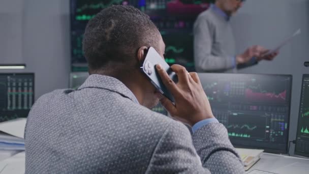 アフリカ系アメリカ人男性の金融アナリストは電話で話します 銀行のオフィスで動作します リアルタイム株式 取引所市場チャートとコンピュータモニタとデジタル画面 — ストック動画