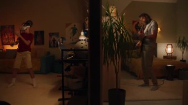 Adam yatak odasında elektro gitar çalıyor. VR kulaklıklı genç çocuk kablosuz kontrolörleri kullanarak arttırılmış realite video oyunu oynuyor. Duvarla ayrılmış iki odanın görüntüsü. Komşuluk ve yaşam tarzı kavramı.