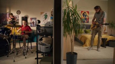 Adam yatak odasında gitar çalıyor. Genç oğlan davul çalar ve genç kız ukulele çalar, yatağa atlar. Duvarla ayrılmış iki oda ya da dairenin görüntüsü. Müzik anlayışı, mahalle ve yaşam tarzı.