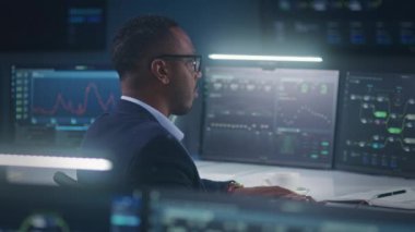 Afrikalı Amerikalı yazılım mühendisi bilgisayar ve veri sunucusu ve engelleme ağı olan büyük dijital ekranların önünde oturuyor, gerçek zamanlı analiz çizelgeleriyle modern izleme odasında çalışıyor..