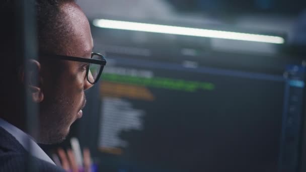 アフリカ系アメリカ人のソフトウェアエンジニアは 表示されたビッグデータサーバーとブロックチェーンネットワークを備えたコンピュータ画面を見て 監視オフィスでリアルタイム分析チャートを監視します サイバーセキュリティの概念 — ストック動画