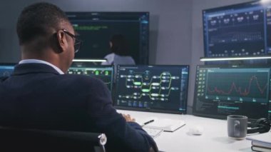 IT teknik uzmanı modern izleme ofisindeki engelleme ağı veritabanına sahip bilgisayarın önünde oturuyor. Yazılım programcıları büyük dijital ekranlarda gerçek zamanlı analiz grafikleriyle çalışırlar..